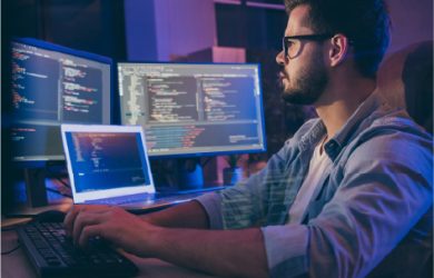 Web developer coding on multiple computer screens at night in modern office